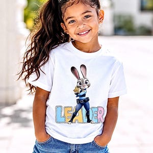 May include: A white t-shirt with a cartoon image of Judy Hopps from Zootropolis, holding a badge. The word "LEADER" is printed below the image in colourful letters. The shirt is worn with denim shorts.