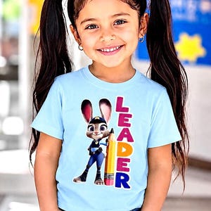 May include: A light blue t-shirt with a cartoon image of Judy Hopps from Zootropolis holding a pencil, and the word "LEADER" in colourful letters. The shirt is worn by a young girl with dark hair in pigtails.