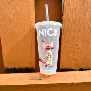 May include: A frosted plastic tumbler with a lid and straw, featuring the name NICK in white capital letters above a graphic of a cartoon fox wearing pink sunglasses, a light-colored short-sleeved shirt, and tan shorts, standing in a confident pose.