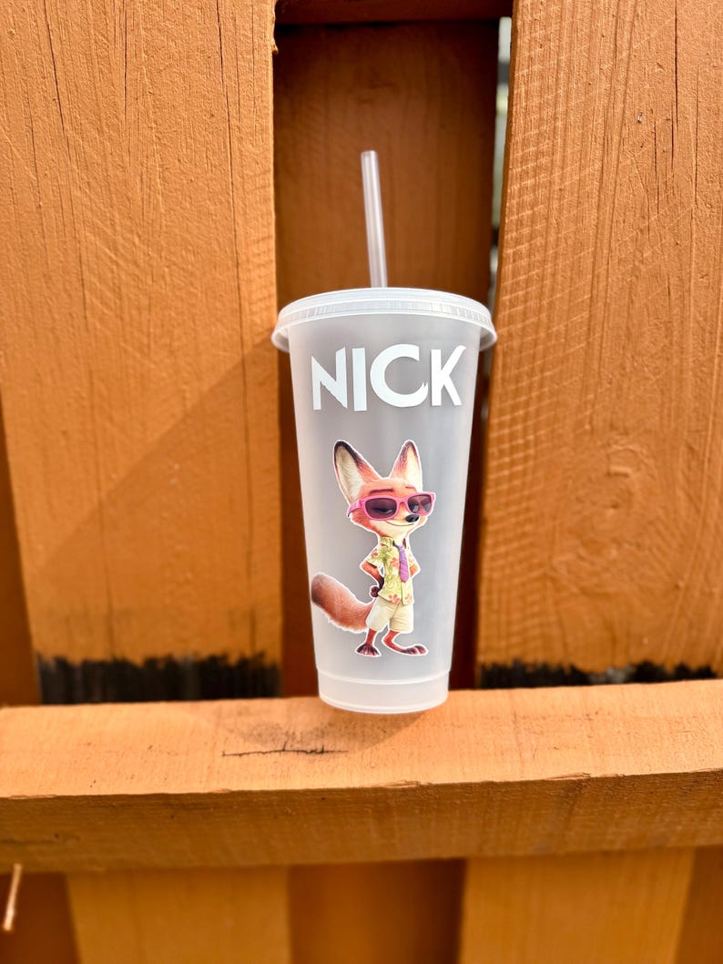 May include: A frosted plastic tumbler with a lid and straw, featuring the name NICK in white capital letters above a graphic of a cartoon fox wearing pink sunglasses, a light-colored short-sleeved shirt, and tan shorts, standing in a confident pose.