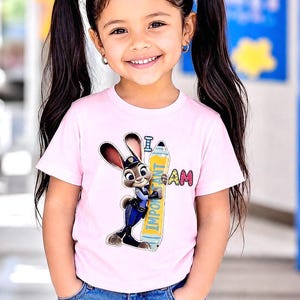 K&ouml;nnte beinhalten: Ein junges M&auml;dchen tr&auml;gt ein hellrosa T-Shirt mit einem Cartoon-Hasen, der einen Bleistift mit der Aufschrift "Important" h&auml;lt. Die Worte "I AM" stehen ebenfalls auf dem Shirt. Das M&auml;dchen l&auml;chelt und tr&auml;gt eine blaue Jeans.