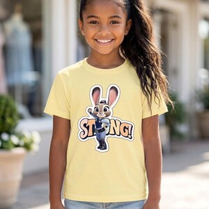 Op de afbeelding: Een lichtgeel T-shirt met een cartoon konijn en de tekst "STRONG!" erop gedrukt. Het T-shirt wordt gedragen door een jong meisje met donker haar en een spijkerbroek.