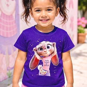 May include: A young child wearing a purple t-shirt with a cartoon bunny graphic and the text "DON'T WORRY BE HOPPY!". The child is also wearing pink shorts and has pink bows in their hair.