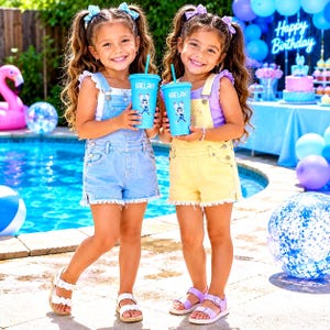 May include: Two children wearing denim dungarees, one in light blue and one in pale yellow, each holding a light blue plastic tumbler with a straw. The tumblers feature a cartoon rabbit graphic and the name Kaelany printed in white text.