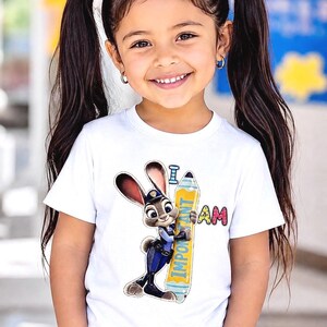 K&ouml;nnte beinhalten: Ein junges M&auml;dchen tr&auml;gt ein wei&szlig;es T-Shirt mit einem Cartoon-Bild eines Hasen-Polizisten, der einen gelben Bleistift mit dem Wort "IMPORTANT" h&auml;lt. Der Text "I AM" steht &uuml;ber dem Bleistift. Das M&auml;dchen hat dunkle Haare in Z&ouml;pfen und l&auml;chelt.