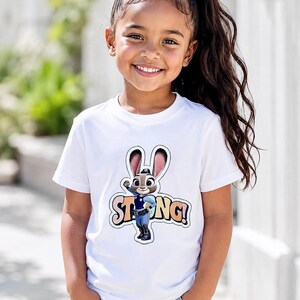 Op de afbeelding: Een wit T-shirt met een cartoonafbeelding van Judy Hopps uit Zootopia, met het woord "STRONG!" in kleurrijke letters. Het T-shirt wordt gedragen door een jong meisje met lang, donker haar en een spijkerbroek.