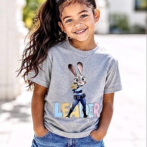 May include: A young girl in a grey t-shirt featuring Judy Hopps from Zootopia, holding a badge. The word "LEADER" is printed below the image in colourful letters. She is also wearing denim shorts.