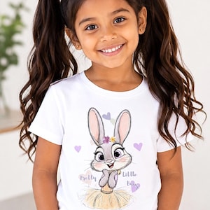 May include: A white short-sleeved t-shirt featuring a graphic illustration of a grey rabbit with large eyes, wearing a yellow skirt and a small flower crown. The text Pretty Little Bunny is printed in purple script alongside small pastel hearts.