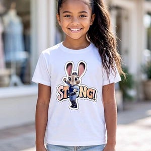 Op de afbeelding: Een wit T-shirt met een cartoon konijn karakter in een politie-uniform en het woord "STRONG!" in oranje en wit. Het T-shirt wordt gedragen door een jong meisje met lang donker haar en een spijkerbroek short.