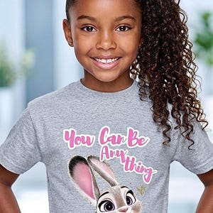 May include: A young girl smiles, wearing a gray t-shirt with the text "You Can Be Anything" in pink script. The shirt features a cartoon image of a bunny in a police uniform. The girl has curly brown hair.