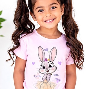 May include: A light pink short-sleeved t-shirt featuring a graphic illustration of a cartoon rabbit wearing a tutu and a flower crown, surrounded by small purple hearts, with the text Pretty Little Bunny printed in a cursive font below the rabbit.