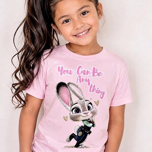 May include: Pink t-shirt featuring a cartoon image of Judy Hopps from Zootopia. The shirt has the text "You Can Be Anything" in pink script. The girl in the image is wearing a pink scrunchie in her hair.
