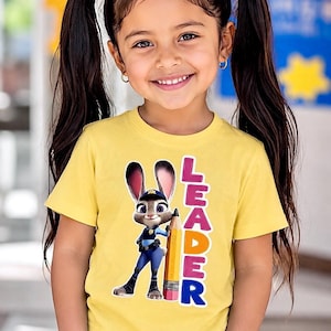 May include: A young girl wearing a yellow t-shirt featuring a cartoon rabbit character holding a pencil. The word "LEADER" is printed vertically on the shirt in pink, orange, and blue letters. The girl is smiling and wearing blue jeans.
