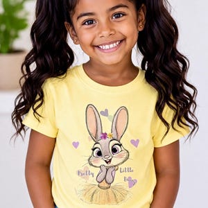 May include: A bright yellow short-sleeved t-shirt featuring a graphic of a cartoon rabbit wearing a yellow tutu and a small flower crown. The rabbit is surrounded by small pastel hearts, with the text Pretty Little Bunny printed in purple.