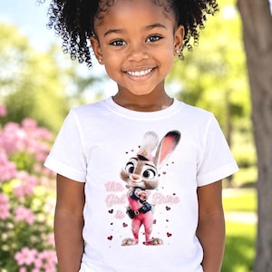 May include: A young girl wearing a white t-shirt featuring a cartoon bunny character in a pink uniform. The shirt has the text "This Girl is Brave" with heart accents. The girl is smiling, with a background of green and pink flowers.