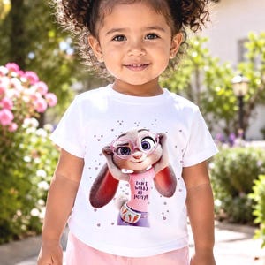 May include: A young child wearing a white t-shirt with a cartoon image of a bunny wearing glasses and the text "DON'T WORRY BE HOPPY!". The child is also wearing pink shorts and has pink bows in their hair.
