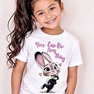 May include: A white t-shirt featuring a cartoon image of a bunny wearing a police uniform, with the text "You Can Be Anything" in pink. The girl is wearing a pink scrunchie in her hair.