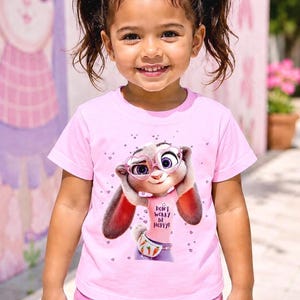 May include: A young girl smiles, wearing a pink t-shirt and shorts. The shirt features a cartoon bunny with glasses and the text "DON'T WORRY BE HOPPY!". The girl has brown hair styled with pink bows.