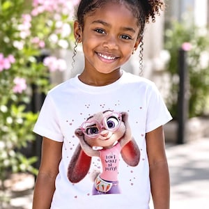 May include: A white t-shirt featuring a cartoon image of Judy Hopps from Zootopia, wearing pink glasses and a fanny pack. The shirt has the text "DON'T WORRY BE HOPPY!". The girl is wearing blue jeans.
