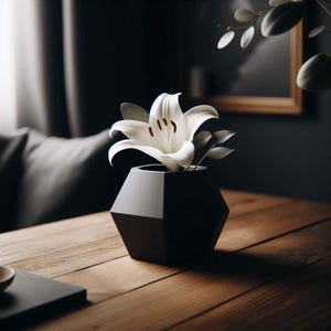 Könnte beinhalten: Eine weiße Lilie mit braunen Staubgefäßen in einer schwarzen, geometrischen Vase. Die Vase steht auf einem Holztisch. Der Hintergrund ist dunkel, mit einem Rahmen und Blättern.