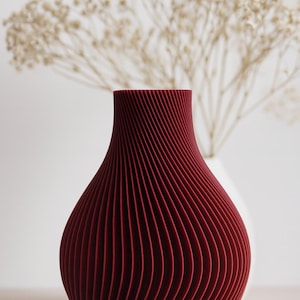 Könnte beinhalten: Eine weinrote Vase mit einem einzigartigen, gerippten Design. Die Vase hat einen breiten, abgerundeten Boden, der sich zu einem schmaleren Hals verjüngt. Im Hintergrund befinden sich eine weiße Vase und Trockenblumen, die eine minimalistische Ästhetik schaffen.