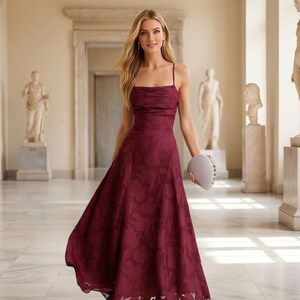 May include: A burgundy-colored formal dress with a floral pattern. The dress features a square neckline, spaghetti straps, and a flowing A-line skirt. A small, light-colored clutch is held in the hand. The dress is worn with silver sandals.