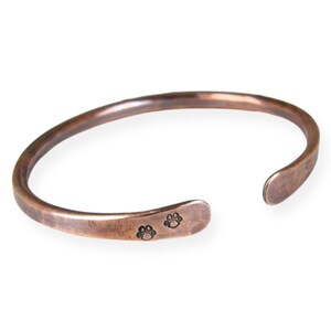May include: A copper cuff bracelet with a hammered finish and two paw print engravings.