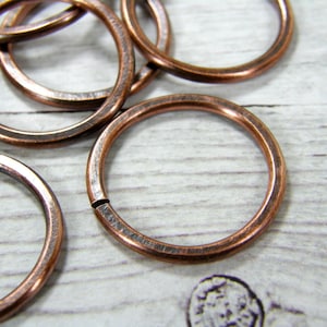 May include: A close-up of several copper jump rings. The rings are smooth and have a slightly textured finish. They are arranged in a random pattern on a white wooden surface.