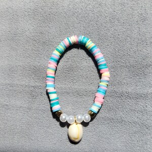 Op de afbeelding: Een kleurrijke armband met pastelkleurige schijfkralen in roze, blauw, geel en wit. De armband is versierd met imitatieparels en een schelpenhanger. De armband is ongeveer 18 cm lang.
