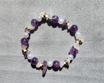 May include: A beaded bracelet featuring amethyst, white pearls, and rose quartz stones. The bracelet has gold-colored accents and is strung on an elastic band. The gemstones are in various shapes and sizes, creating a unique and elegant accessory.