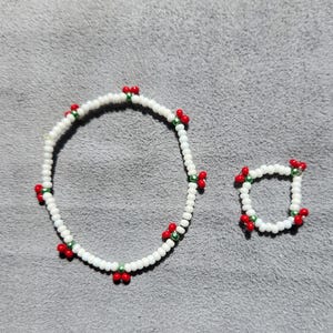 May include: White beaded bracelet and ring set. The bracelet and ring are adorned with red and green accents. The jewelry is set against a gray background.