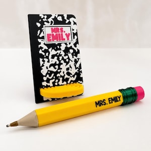 May include: A black and white composition notebook-style phone stand with a yellow shelf and the text MRS. EMILY in pink, paired with a yellow pencil-shaped pen featuring a green ferrule, pink eraser, and the black text MRS. EMILY.