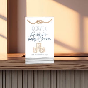 May include: A white sign with the text "Decorate a block for baby Brown" in a script font. The sign also features three toy blocks with the letters A, B, and C. A rope design is at the top. The sign is on a wooden surface.