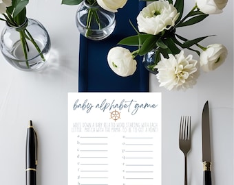 May include: A baby shower game with the text "baby alphabet game" and a nautical wheel graphic. The game is on a white card with a navy blue runner, surrounded by white flowers in glass vases, a pen, and silverware.