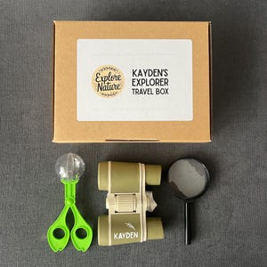 May include: A brown cardboard box labelled "Kayden's Explorer Travel Box" with a green bug catcher, olive green binoculars, and a magnifying glass. The box has a white label with the words "Explore Nature".