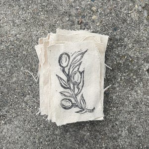 May include: A stack of off-white fabric patches with a black botanical print. The print features a stylized floral design with leaves and oval shapes. The patches have raw, unfinished edges and are arranged on a gray concrete surface.