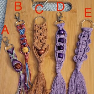 May include: A collection of handmade macrame keychains in various designs and colors. Keychains feature wooden beads, tassels, and metal clasps. The keychains are displayed on a wooden cutting board.