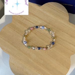 Puede incluir: Una pulsera de cuentas con cuentas transparentes, azules, rosas, verdes y blancas. Algunas cuentas tienen un diseño floral azul y blanco. La pulsera está sobre una bandeja de madera clara en forma de flor. El logo "Shelbz Randomspired" es visible.