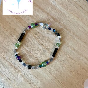 Puede incluir: Una pulsera de cuentas con una mezcla de cuentas coloridas, incluyendo verdes, moradas, negras y transparentes. La pulsera está sobre una superficie de madera. El texto "Shelbz Random-inspired" está en la esquina superior izquierda.