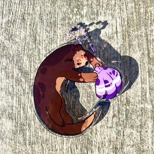 May include: A clear acrylic keychain featuring a cartoon otter holding a purple seashell. The otter is brown with dark brown spots and a light brown belly. The keychain has a purple chain.