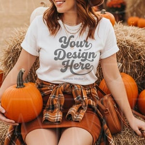 Puede incluir: Una mujer con una camiseta blanca con el texto "Your Design Here" y un sombrero marrón, sentada en una paca de heno rodeada de calabazas. Lleva una falda marrón y una camisa a cuadros atada a la cintura. Un bolso marrón está en su hombro.