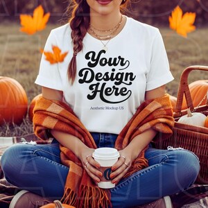 Puede incluir: Una camiseta blanca con el texto "Your Design Here" se usa con jeans azules y una bufanda naranja. La escena incluye calabazas, una cesta de picnic y hojas de otoño. Se sostiene una taza de café en las manos.