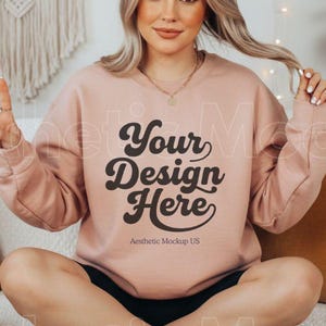 May include: A light pink sweatshirt with the text "Your Design Here" in a retro font. The model is sitting on a bed with pillows and a woven tray with a mug. The sweatshirt is a mock-up for a design.