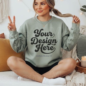Pode incluir: Camisola verde sálvia com o texto "Your Design Here" em uma fonte retrô. A camisola é de gola redonda. A modelo está sentada em uma cama com almofadas e uma bandeja decorativa com uma vela.
