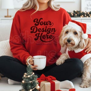 May include: A red sweatshirt with the text "Your Design Here" is worn by a person sitting on a white couch. A small dog in a Santa suit and hat sits next to the person. Presents wrapped in brown paper with red bows and a small Christmas tree are in front.