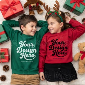以下が含まれることがあります： 「Your Design Here」の文字が入った緑と赤のスウェットを着た2人の子供。白い表面に横たわり、プレゼント、テディベア、松ぼっくりに囲まれています。スウェットはホリデーをテーマにしたコレクションの一部です。
