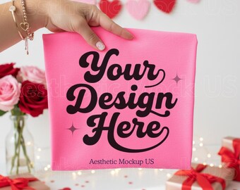 Puede incluir: Una tela cuadrada rosa con el texto "Your Design Here" en negro, con una fuente retro. Las palabras "Aesthetic Mockup US" están debajo del texto principal. La tela se presenta frente a un fondo blanco con decoraciones de San Valentín.