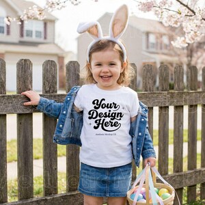 May include: A young child wearing a white t-shirt with the text "Your Design Here", a denim jacket, and a denim skirt. The child is wearing bunny ears and holding a basket with Easter eggs. The child is standing next to a wooden fence.