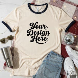 May include: A cream-coloured ringer t-shirt with navy blue trim and the text "Your Design Here" in black script. The shirt is styled with light wash jeans, white trainers, and sunglasses, suggesting a casual style.