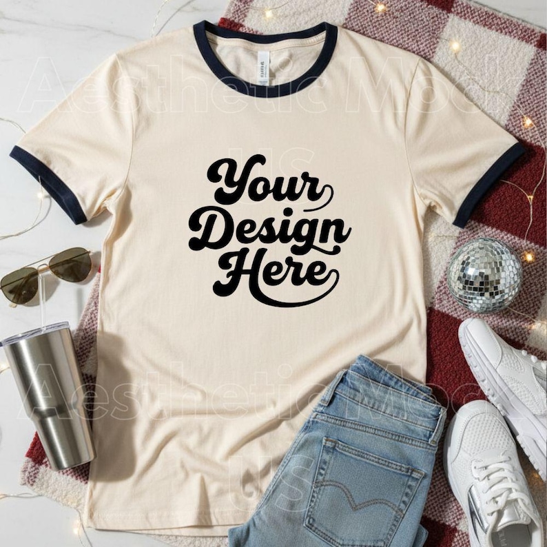 May include: A cream-coloured ringer t-shirt with navy blue trim and the text "Your Design Here" in black script. The shirt is styled with light wash jeans, white trainers, and sunglasses, suggesting a casual style.